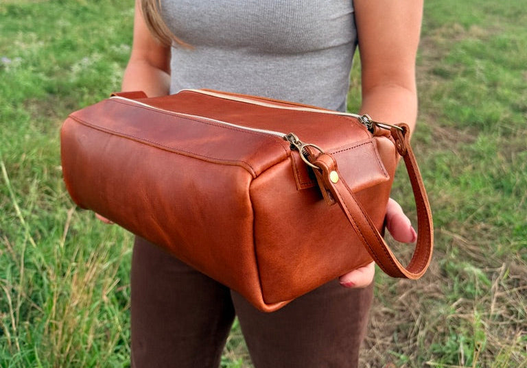 Indigo Laine Road Kit in warm brown leather with double top zippers and a leather side handle, held outdoors.