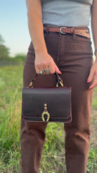 A person outdoors in a grassy field holding a dark brown leather handbag with gold-tone hardware and a horseshoe-shaped clasp.